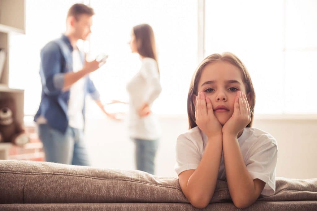 Sad little girl is looking at camera while her parents are arguing in the background