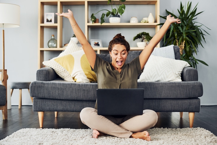 Shot of a young woman cheering while using a laptop on the living room floor at home